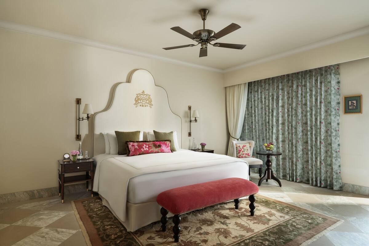 Une chambre avec un grand lit, un ventilateur de plafond, une table de chevet avec une lampe, une petite table avec un vase et une fenêtre avec des rideaux.