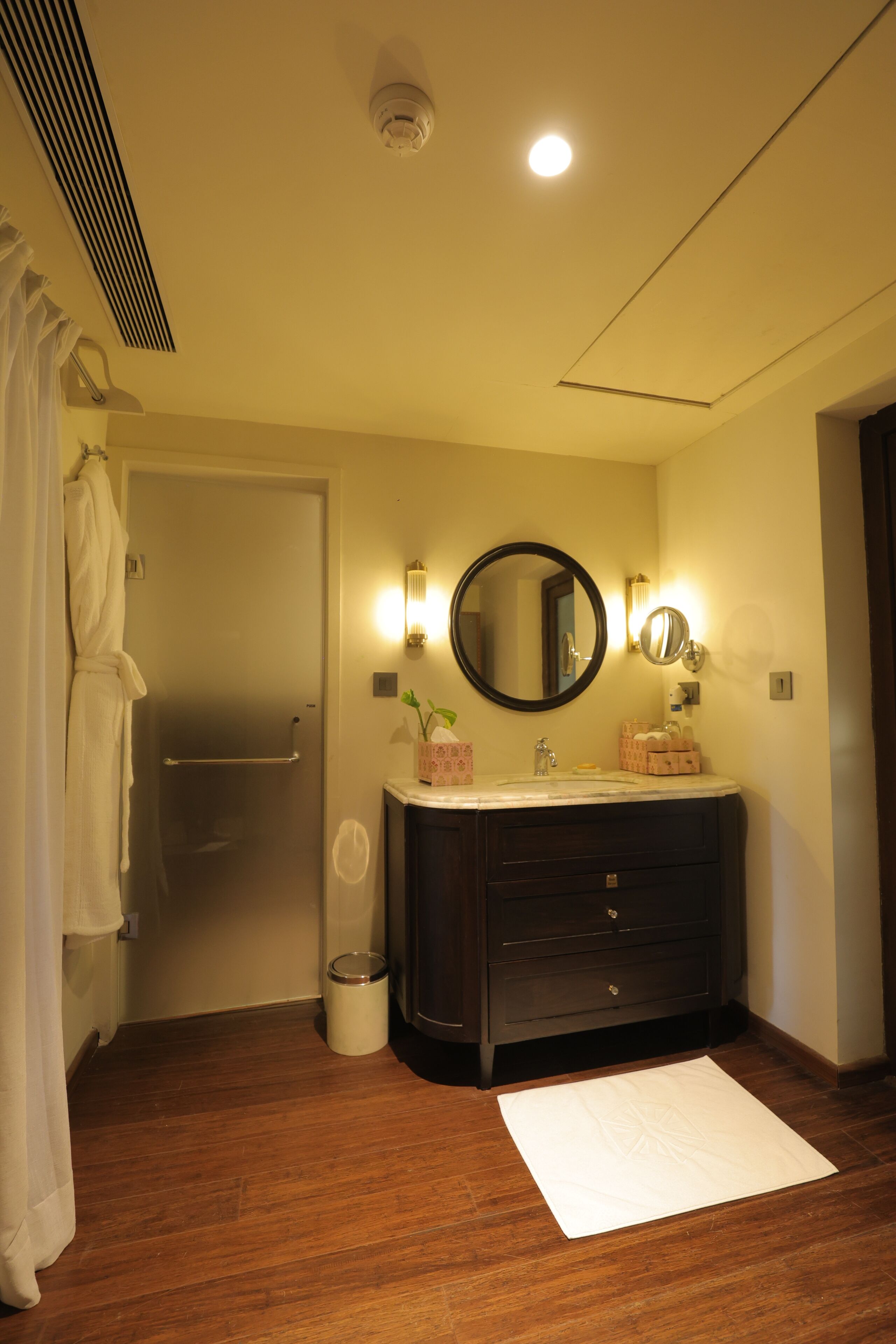 A bathroom with a wooden floor, a white bath mat, a dark wooden vanity, a round mirror, and a towel rack.