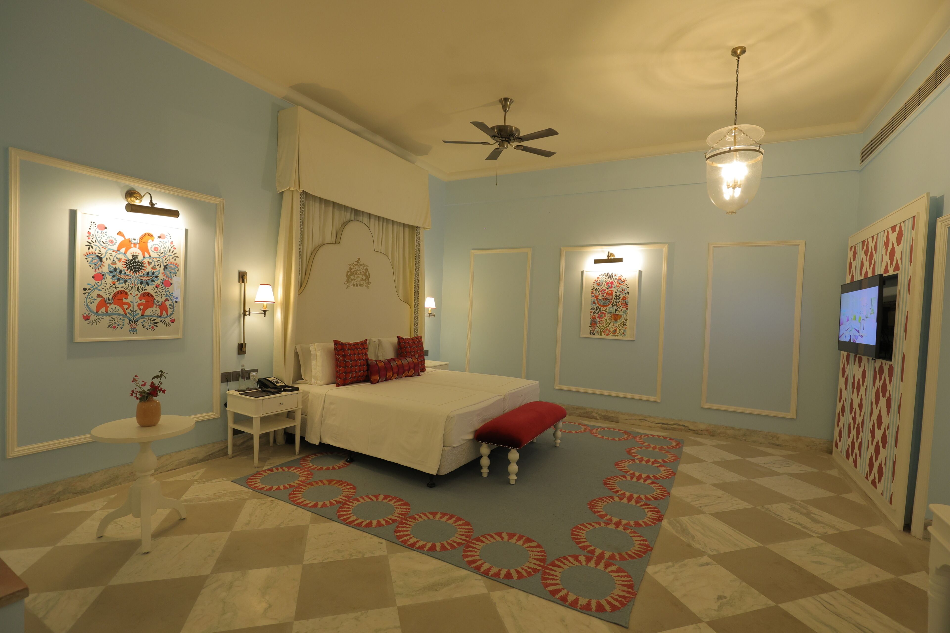Un dormitorio con cama, mesita de noche, banco rojo, ventilador de techo, televisor y alfombra estampada.