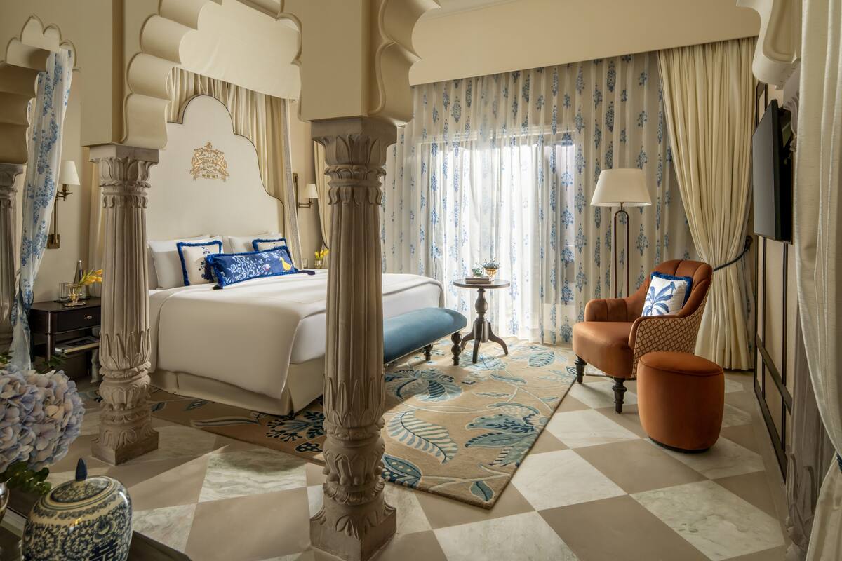 Une chambre d’hôtel avec un grand lit, un coin salon comprenant un fauteuil et un pouf, une petite table et un vase décoratif.