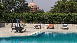 A swimming pool with lounge chairs and a table, surrounded by greenery and a large building in the background. - Clarks Avadh, Lucknow (Lucknow)