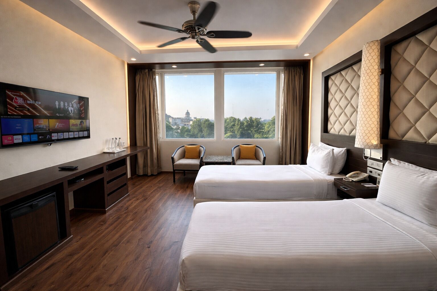 A hotel room with two beds, a TV, a desk, and a view of the city.