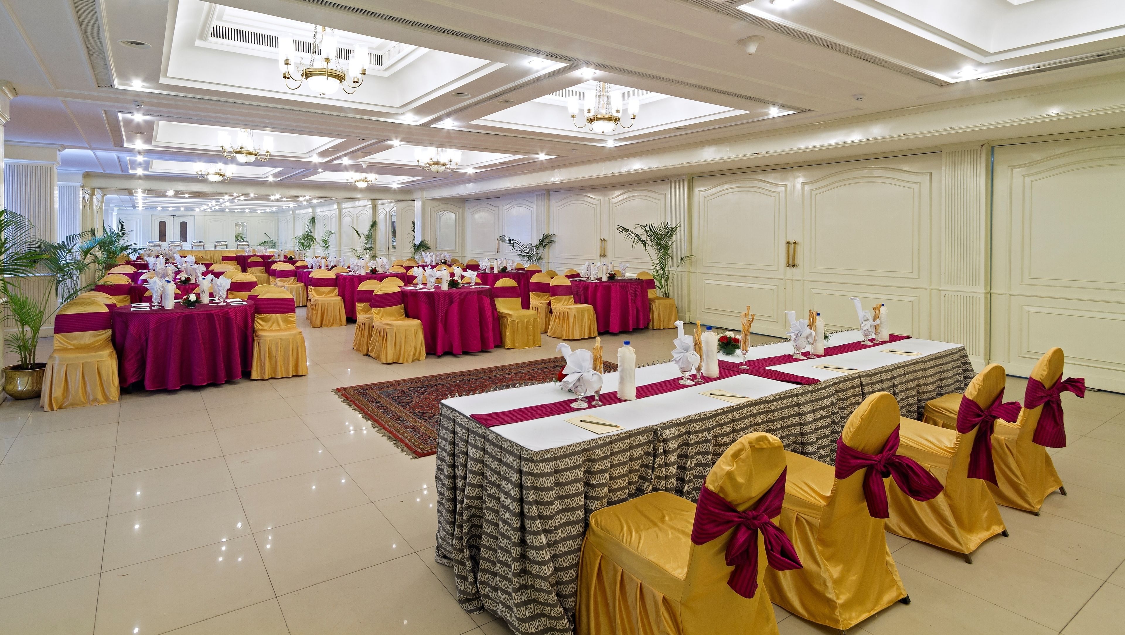 A banquet hall with round tables covered in white tablecloths, yellow chairs with red sashes, and chandeliers on the ceiling.