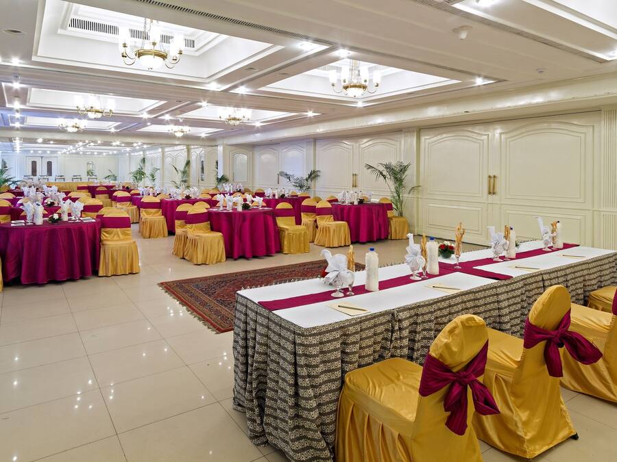 A banquet hall with round tables covered in white tablecloths, yellow chairs with red sashes, and chandeliers on the ceiling.