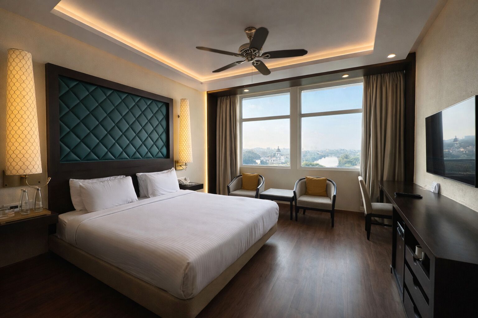 Habitación de hotel con una cama grande, un escritorio, una silla, televisión y ventilador de techo, con vista a un paisaje urbano.