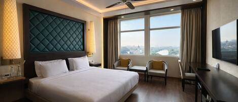 A hotel room with a large bed, a desk, a chair, a TV, a ceiling fan, and a view of a cityscape.