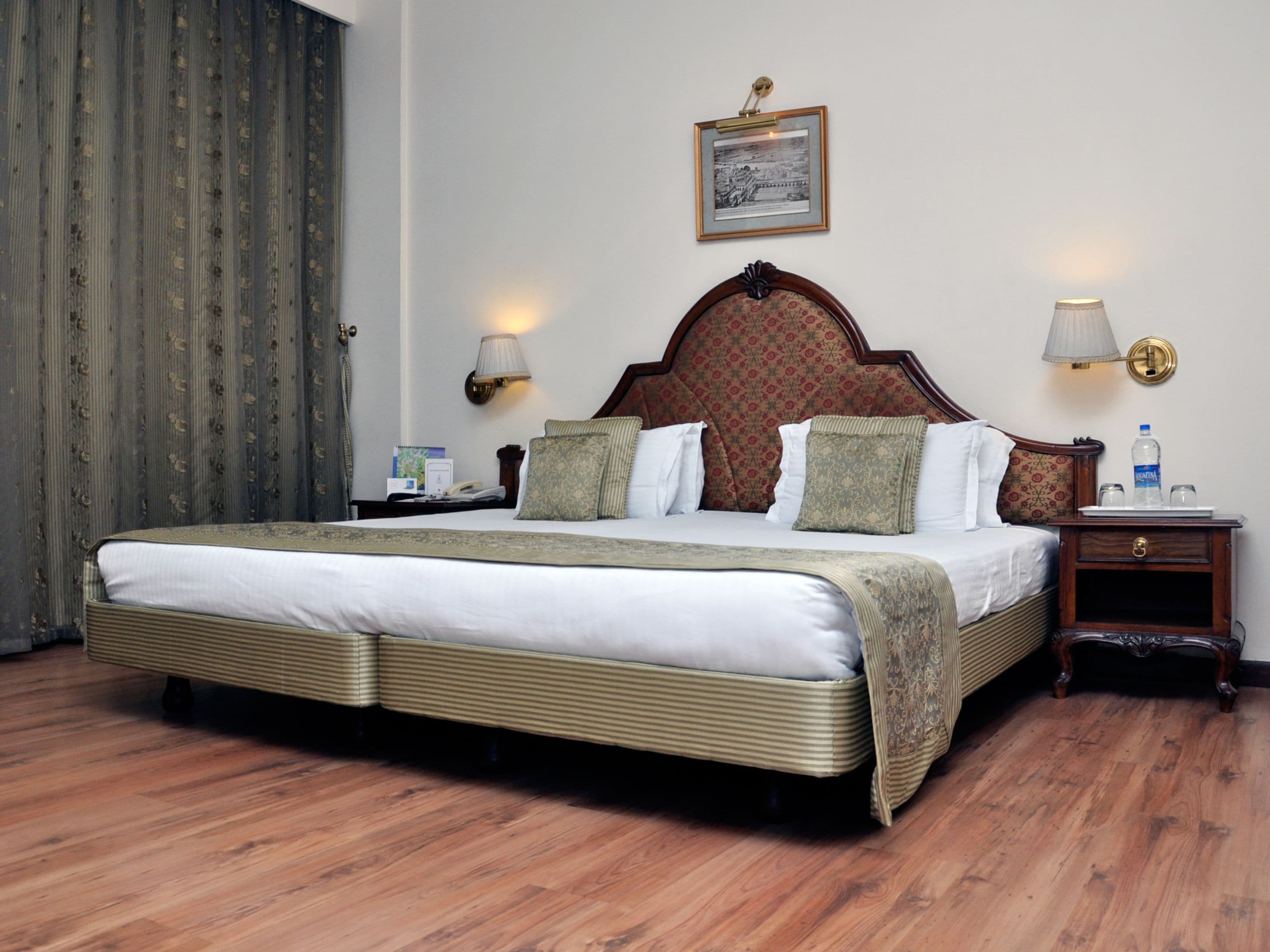 A neatly made bed with a wooden headboard, two bedside tables with lamps, a framed picture on the wall, and a curtain on the left.