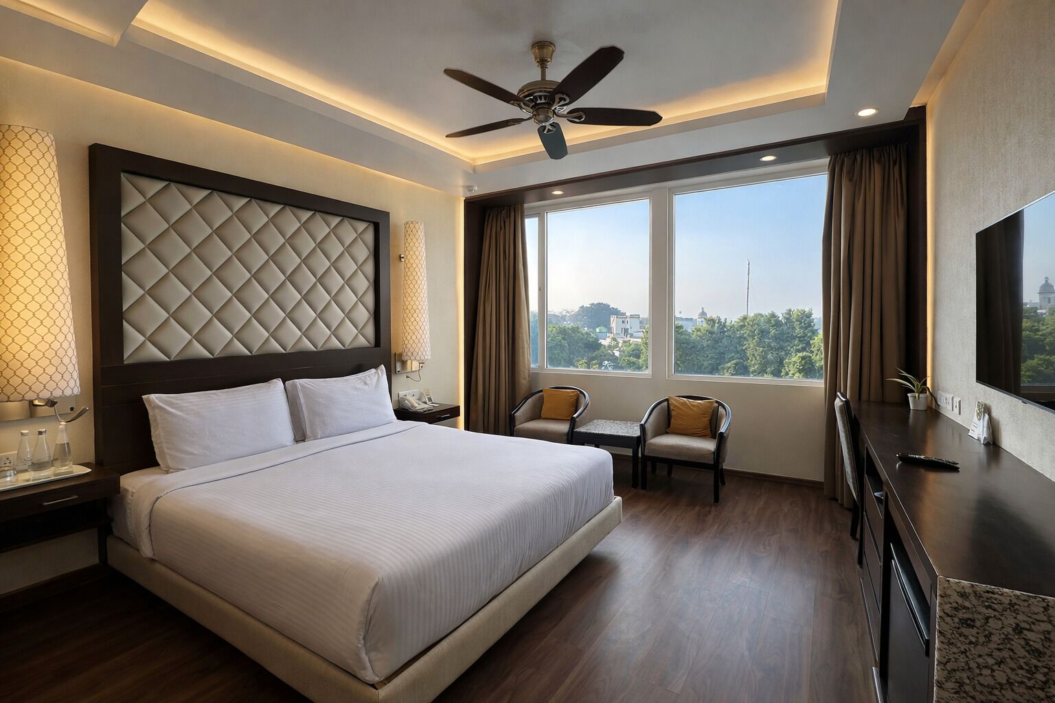 Una habitación de hotel con una cama grande, ventilador de techo, escritorio, dos sillas y un ventanal con vistas.