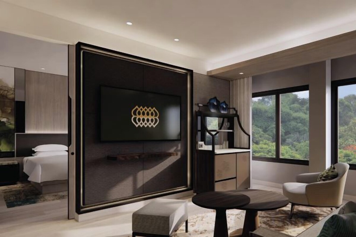 A modern hotel room with a large flat-screen TV mounted on a dark wall, a comfortable seating area with a round table, and a large window offering a view of the outdoors.