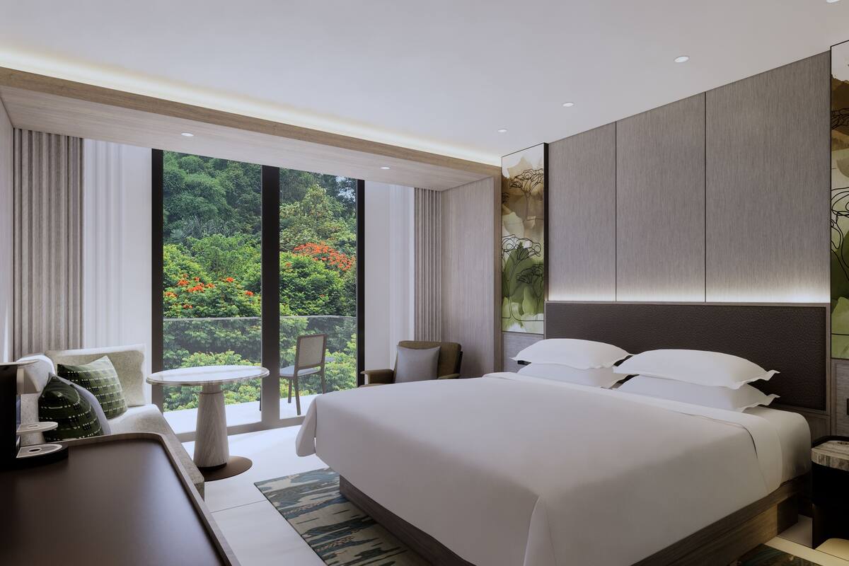 A modern hotel room with a large bed, a desk, a chair, and a view of greenery through the window.