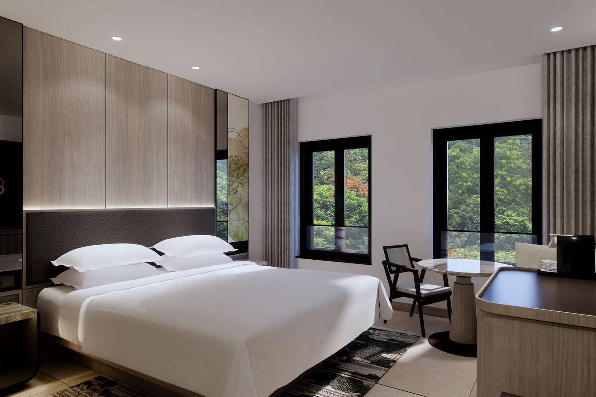 A modern hotel room with a large bed, a desk, and a view of greenery through the windows.