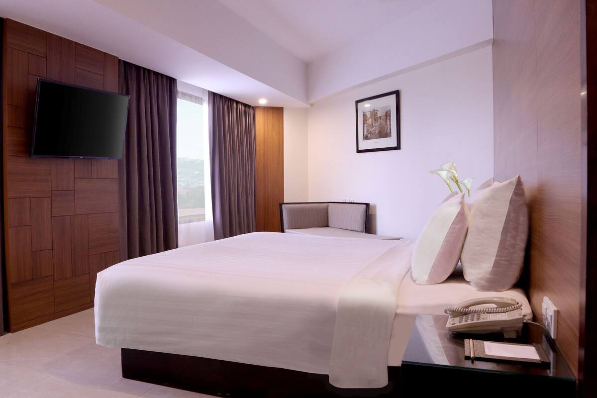 A modern hotel room with a large bed, a flat-screen TV mounted on the wall, a bedside table with a phone and a notepad, and a window with curtains.