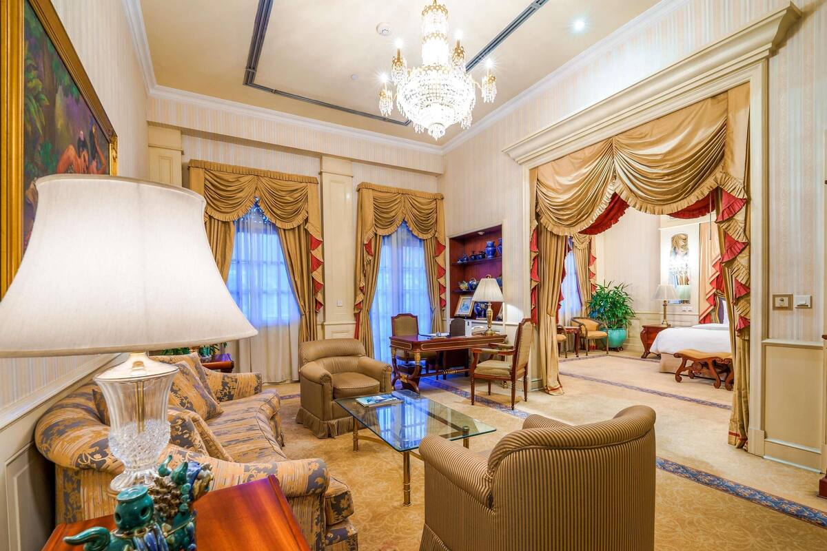 Ein luxuriöses Hotelzimmer mit einem großen Chandelier, aufwendigen Vorhängen, einem bequemen Sofa, einem Glastisch und einem traditionellen Holznachttisch.