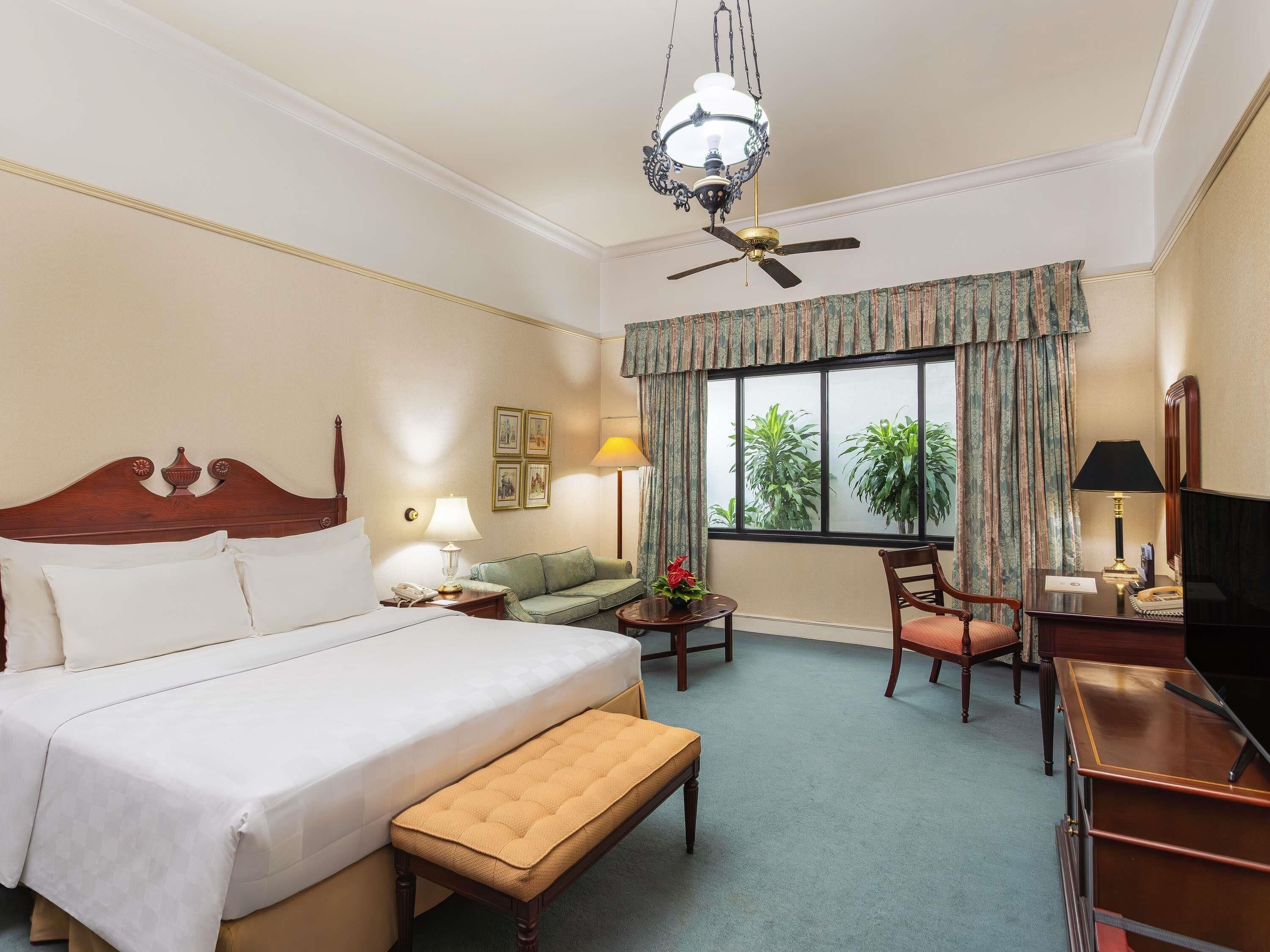 A hotel room with a bed, a desk, a chair, a small table, a ceiling fan, and a window with curtains.