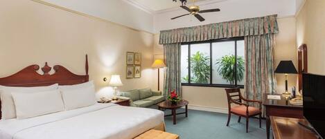 A hotel room with a bed, a desk, a chair, a small table, a ceiling fan, and a window with curtains.