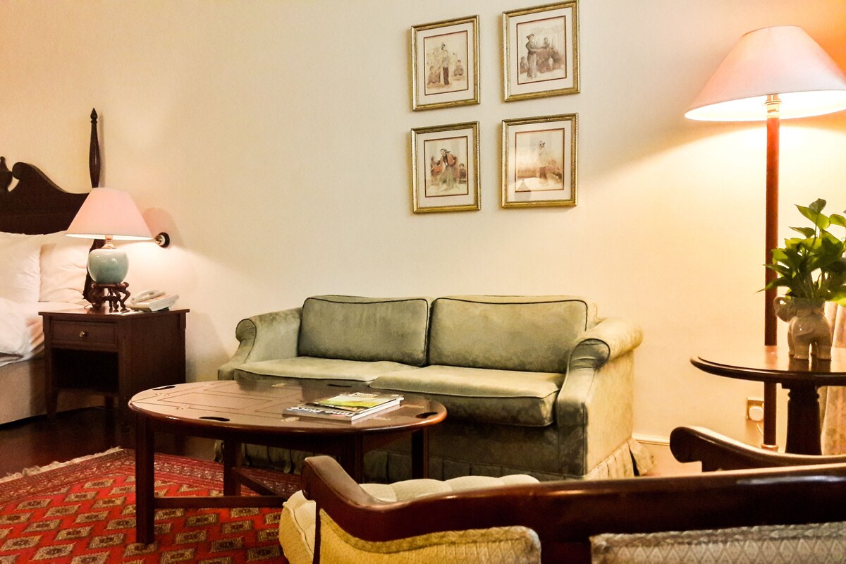 Une chambre d’hôtel avec un lit, un canapé vert, une petite table, une lampe et des tableaux encadrés au mur.