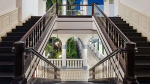 Uma imponente escadaria com dois lances, paredes brancas e vista para o verde através de arcos.