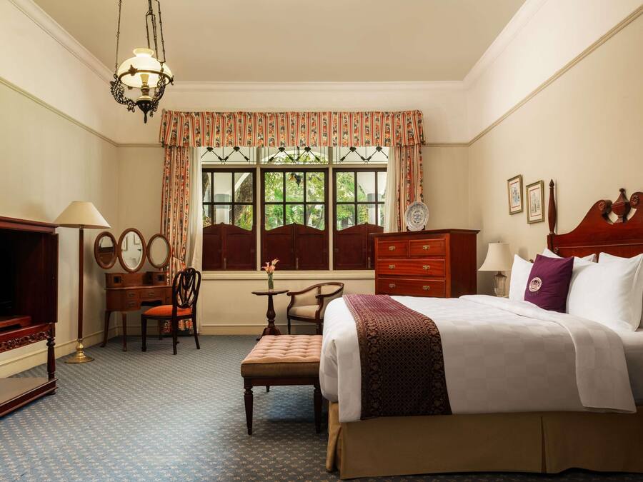 A hotel room with a bed, a desk with a lamp, a chair, a dresser, and a window with curtains.