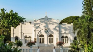 Um edifício branco com janelas em arco e um relógio, rodeado por vegetação exuberante.