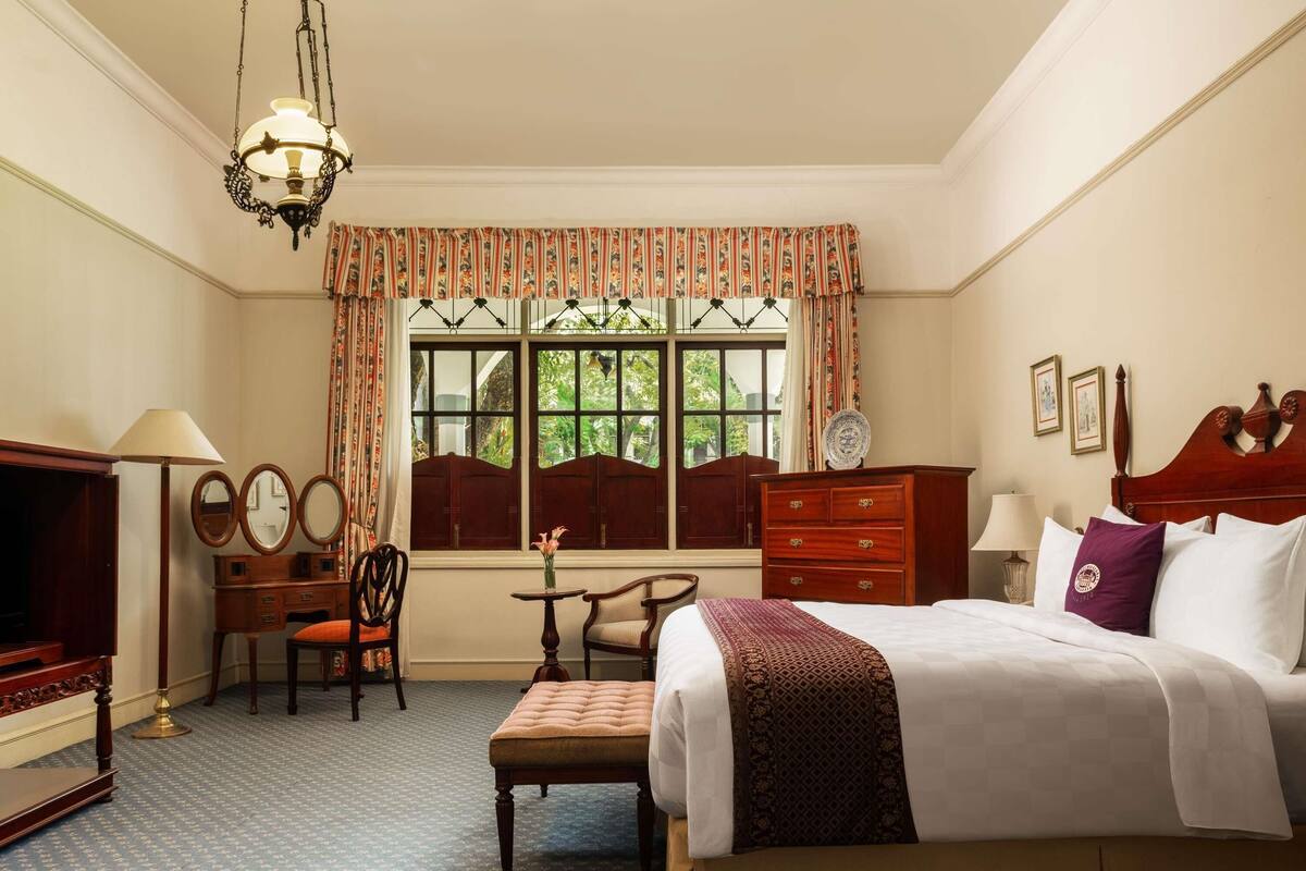 Ein Hotelzimmer mit Bett, Schreibtisch mit Lampe, Stuhl, Kommode und einem Fenster mit Vorhängen.