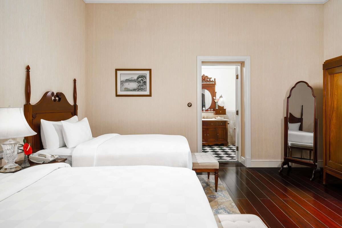Ein Hotelzimmer mit zwei Betten, einem Holzkopfstück, einem gerahmten Bild an der Wand, einem durchgehenden Türspalt erkennbaren Badezimmer und einem Schachbrettfußboden.