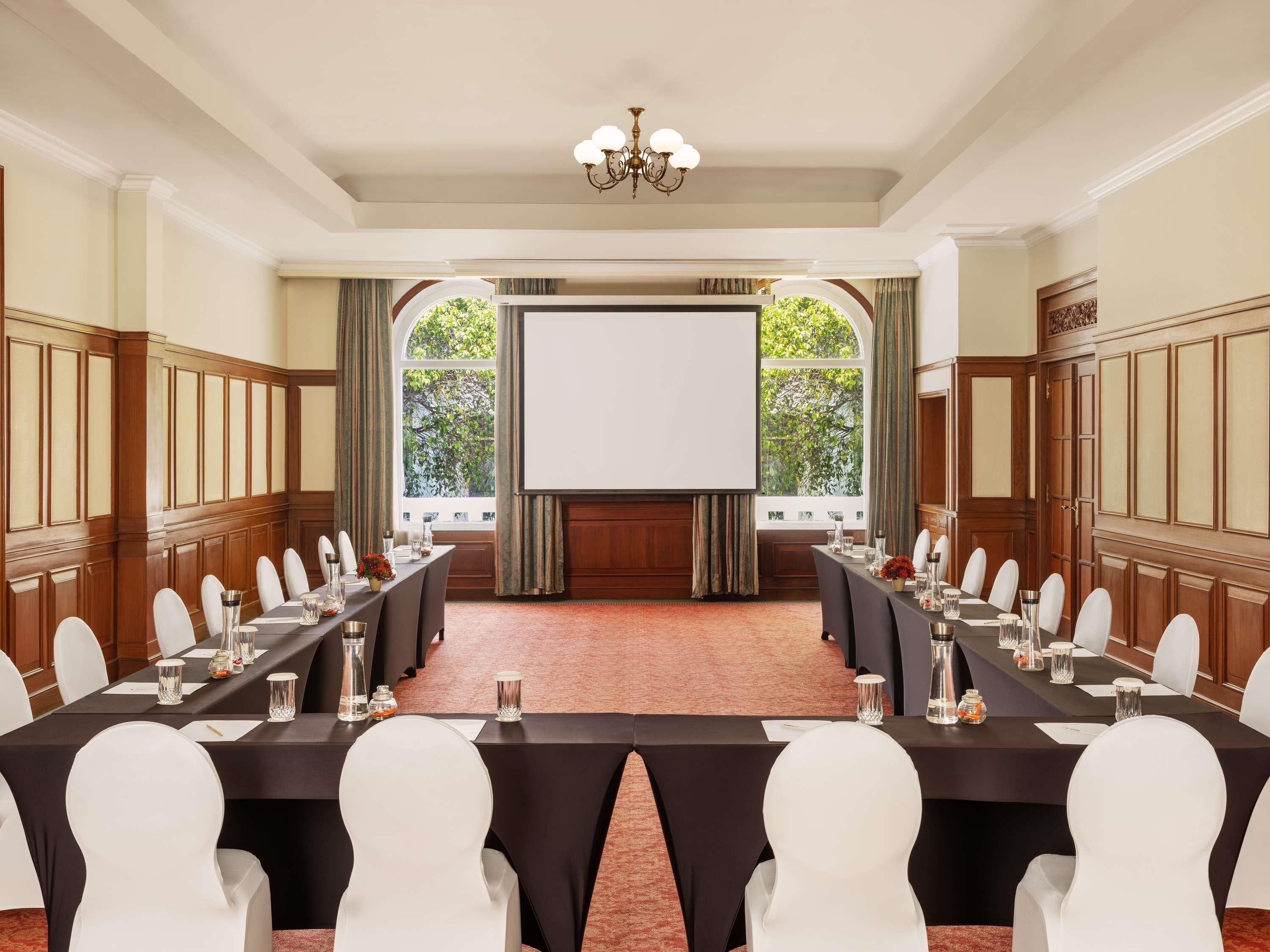 Un salón de conferencias con una pantalla grande, mesas largas y sillas blancas.