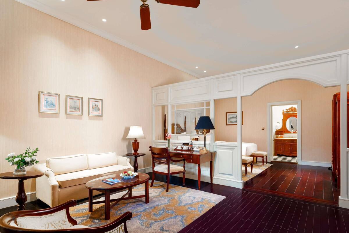 A spacious living room with a sofa, chairs, a coffee table, and a desk. There is a ceiling fan, framed artwork on the walls, and a patterned rug.
