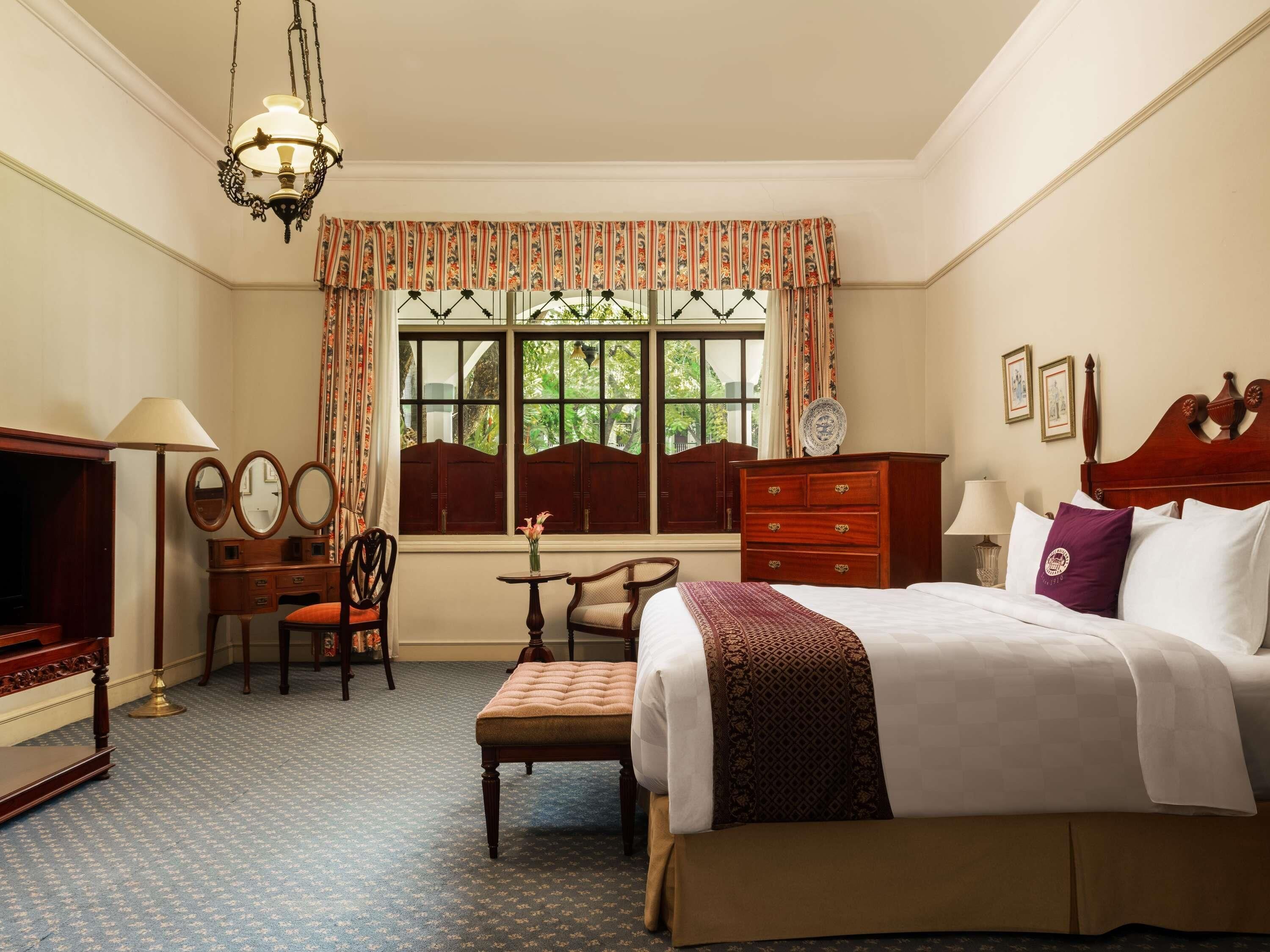 Une chambre d’hôtel avec un lit, un bureau avec une lampe, une chaise, une commode et une fenêtre avec des rideaux.