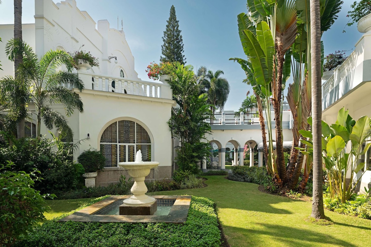 A courtyard with a fountain, lush greenery, and a white building with arched windows and balconies.