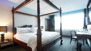 A four-poster bed with a wooden frame and white bedding, a bedside table with a lamp, a chair, and a small table with a glass on it.