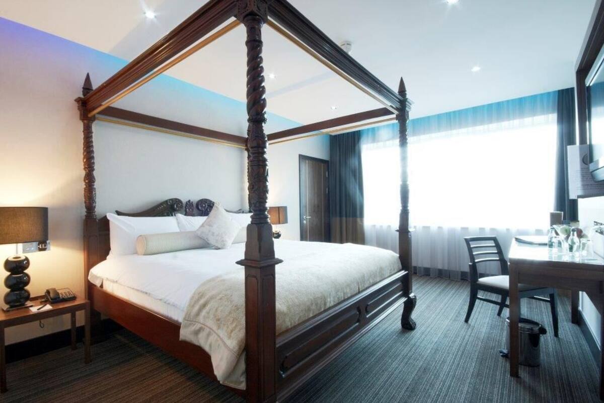 Una cama con dosel de madera y ropa de cama blanca, una mesita de noche con una lámpara, una silla y una pequeña mesa con un vaso encima.