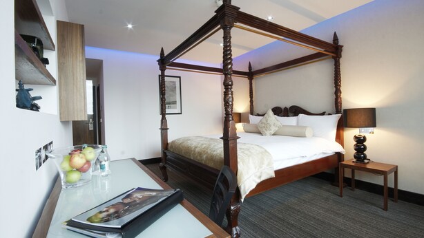 A four-poster bed with a wooden frame, a bedside table with a lamp, and a glass vase with apples on a desk.