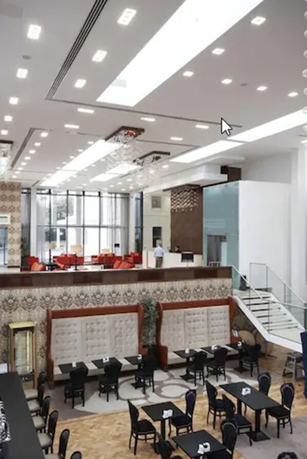 A spacious hotel lobby with a high ceiling, multiple seating areas, and a staircase leading to an upper level.