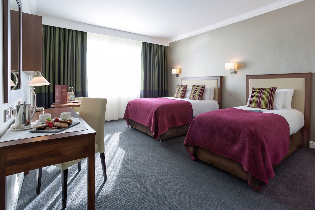 A hotel room with two beds, a desk with a coffee maker, a chair, and a view of the outdoors.