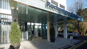 La façade extérieure de l’hôtel Rochester Park, avec son entrée en verre, ses plantes en pot et un ciel dégagé.