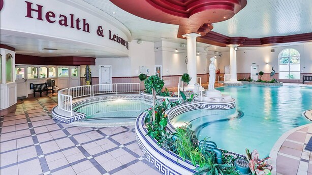 Une grande piscine intérieure avec un sol à carreaux, entourée d’un bord décoratif et de plantes.