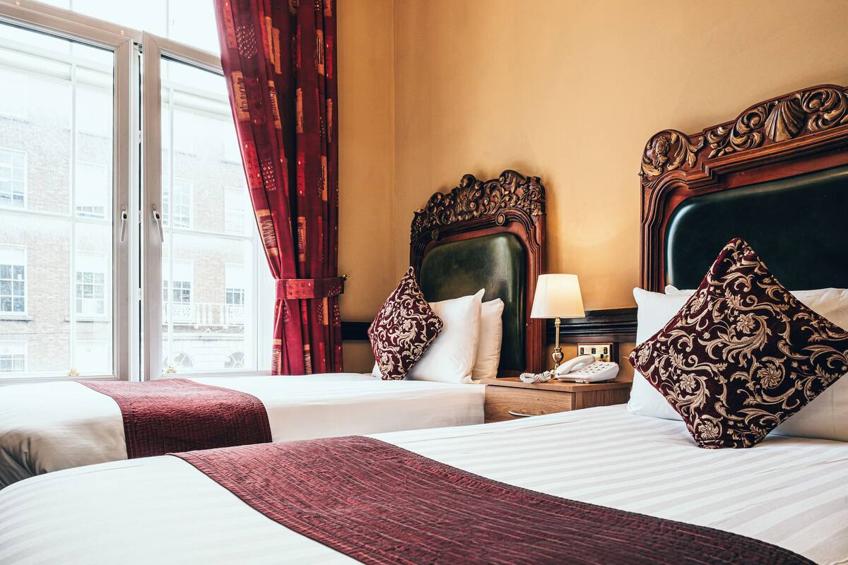 A hotel room with two beds, a nightstand with a telephone, and a window with red curtains.