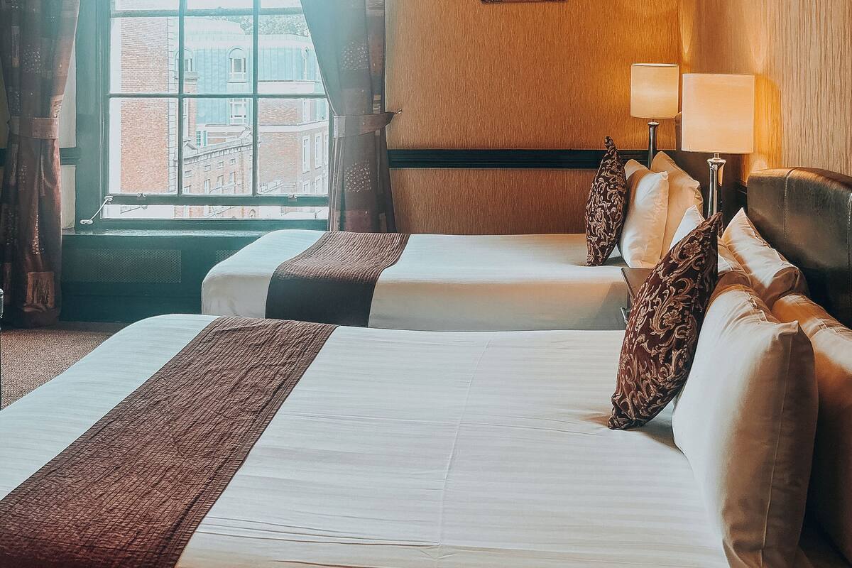 A hotel room with a large bed, two bedside tables, a chandelier, a window with curtains, and a view of buildings outside.