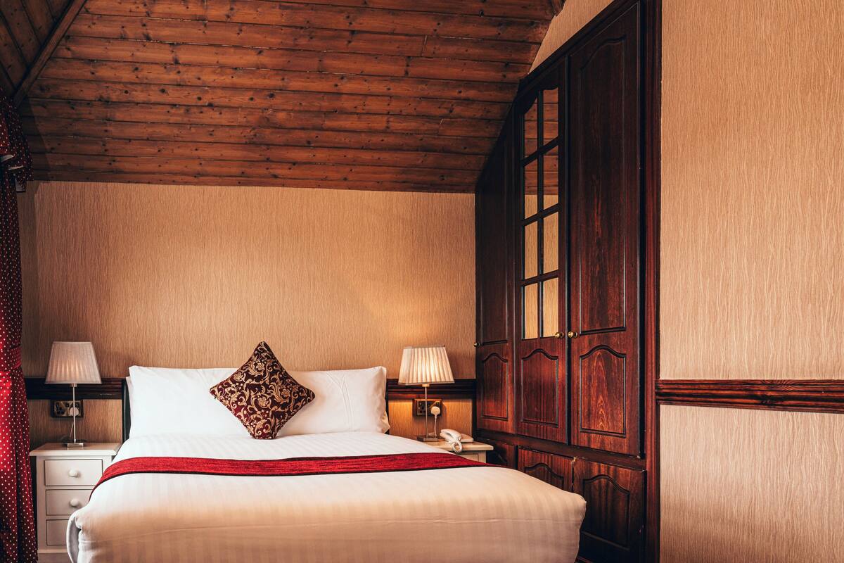 A hotel room with a wooden ceiling, a large bed with white linens, two bedside lamps, a telephone, and a large wooden wardrobe.