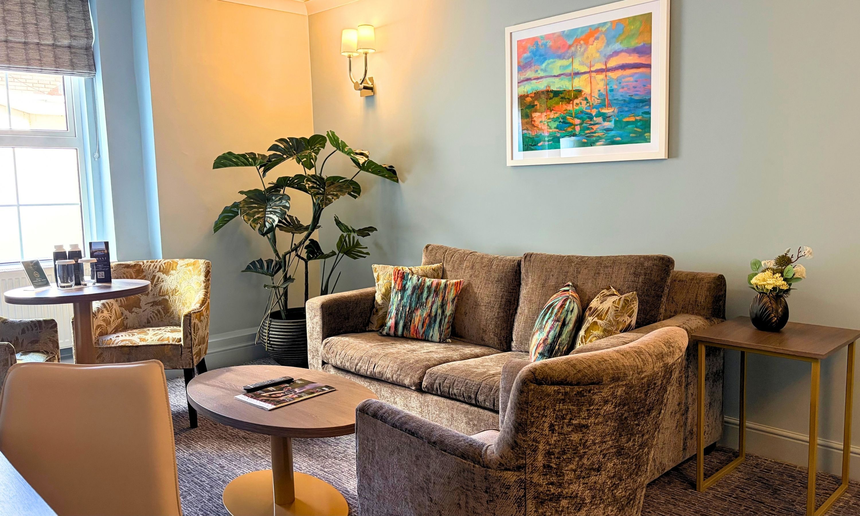 Un salon avec un canapé marron, deux fauteuils, une table basse, une plante en pot, un tableau et une petite table sur laquelle est posé un vase.