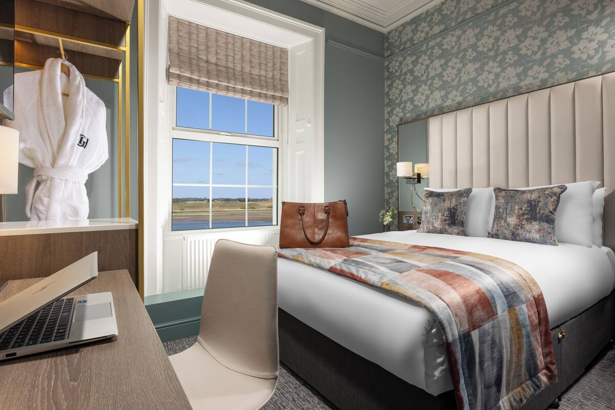 A hotel room with a bed, a desk with a laptop, a chair, a robe hanging, and a window with a view.