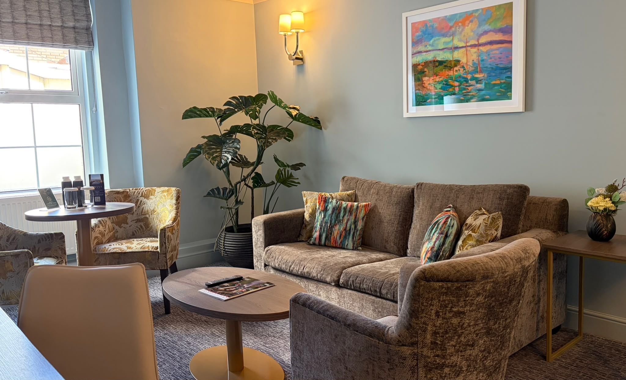 A living room with a sofa, armchairs, a coffee table, a potted plant, a painting, and a wall-mounted lamp.