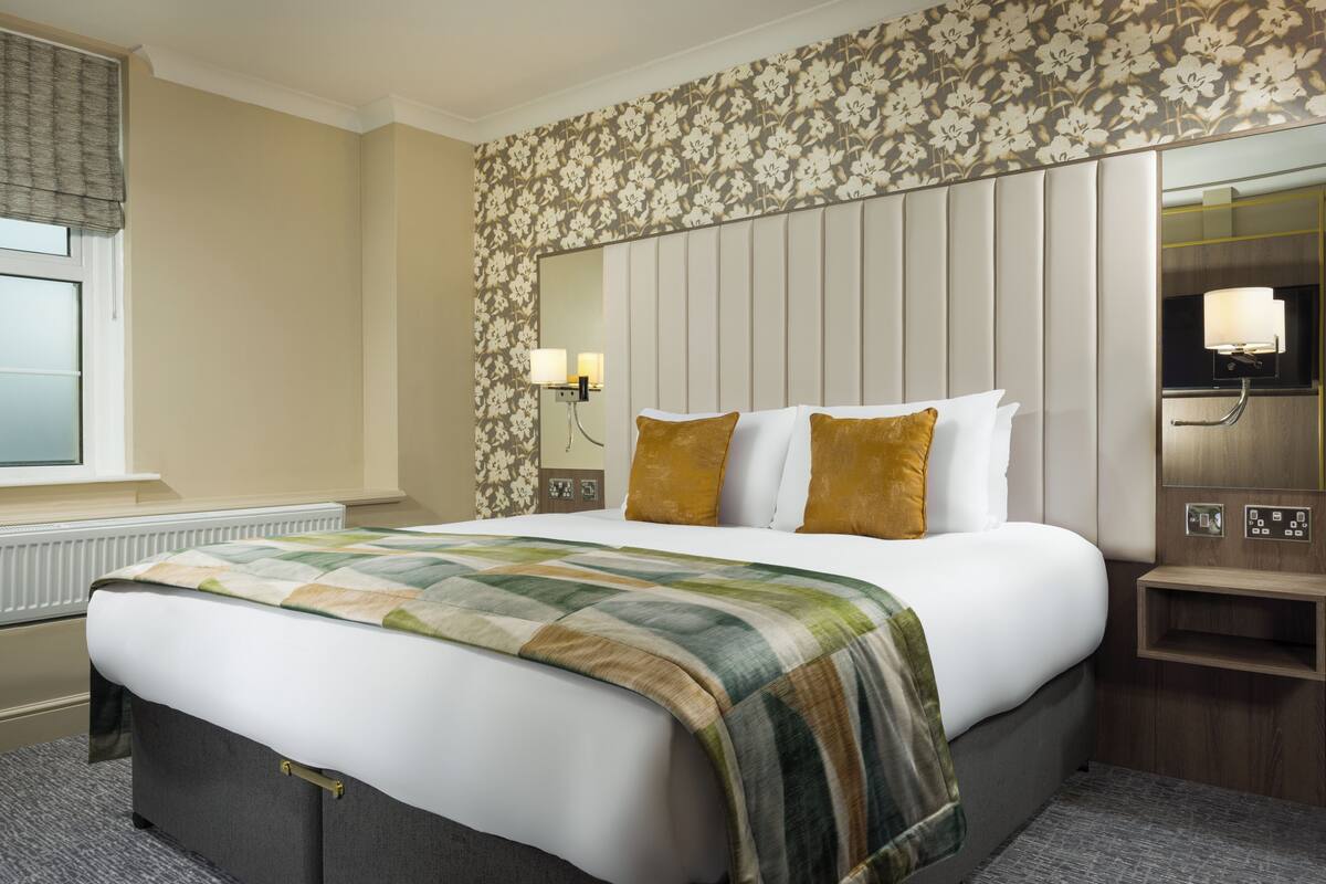 A hotel room with a large bed, a nightstand with a lamp, a wall-mounted mirror, and a floral-patterned wallpaper.