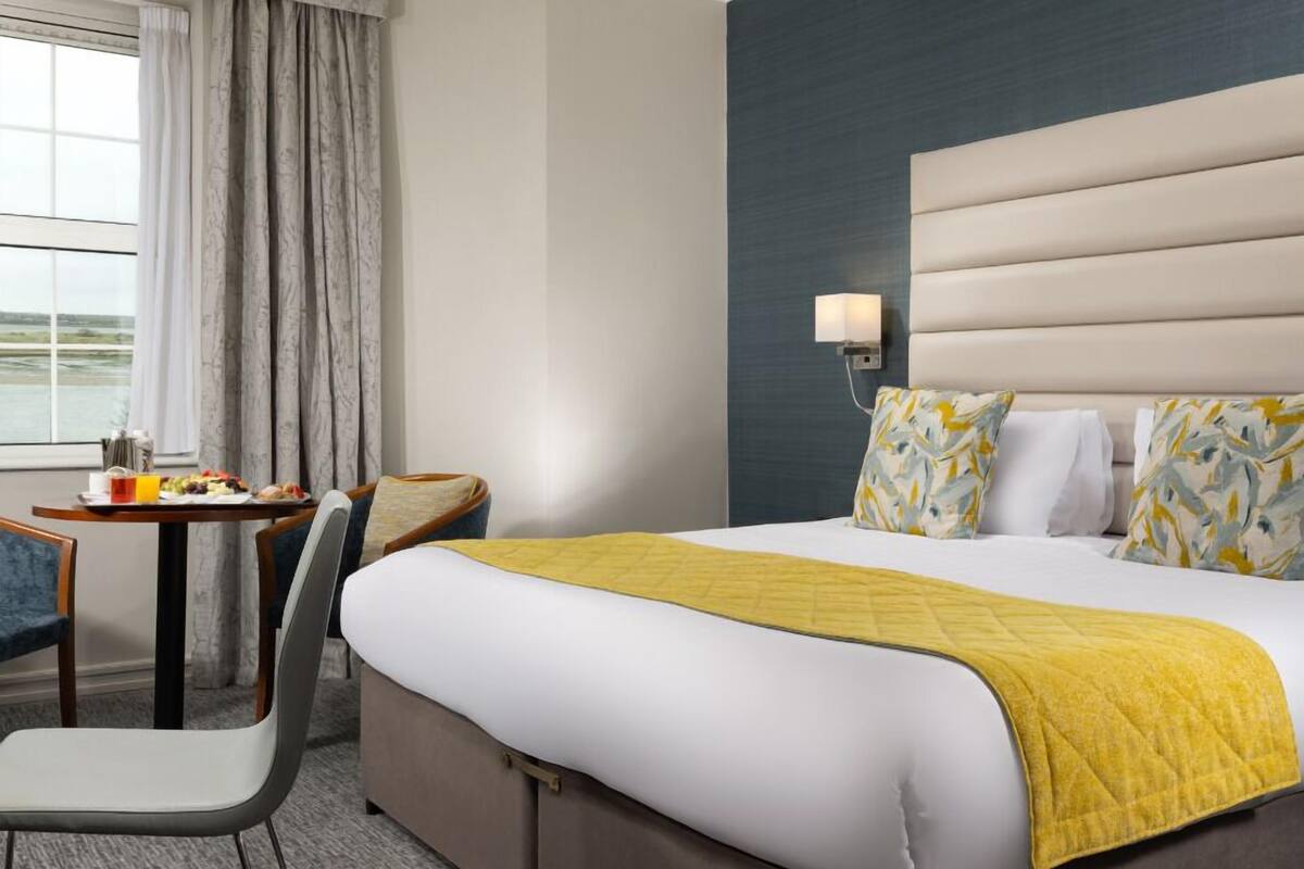 A hotel room with a bed, a desk with breakfast, a chair, and a view of the outdoors.