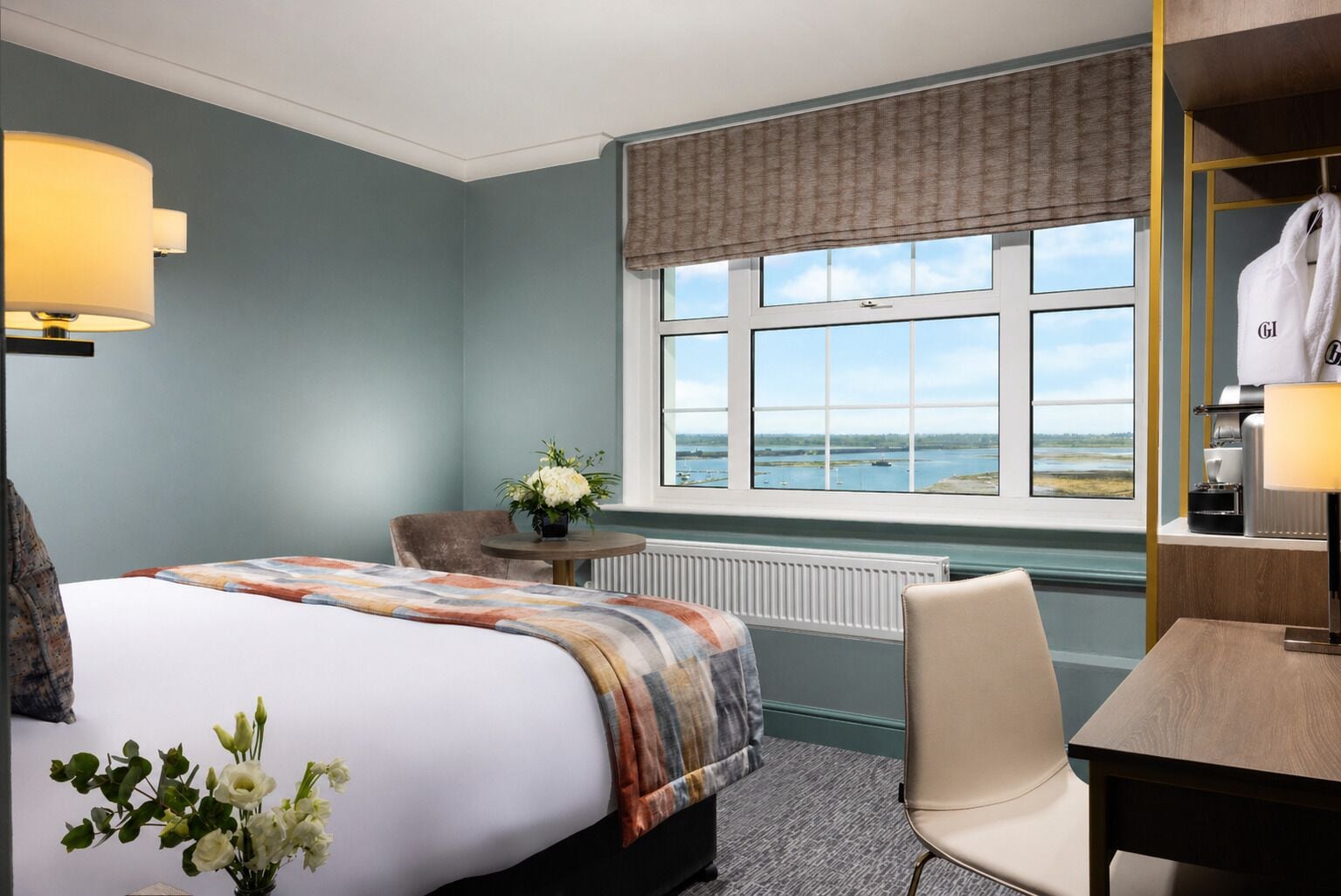 Hotelkamer met een bed, bureau, stoel, lamp, radiator, raam met uitzicht op zee en een tafeltje met lamp.