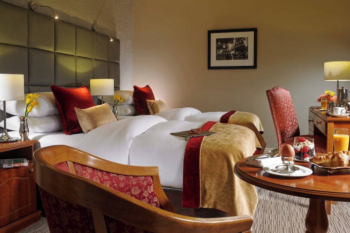 Habitación de hotel con una cama grande, una silla de madera, una mesa de comedor con alimentos para el desayuno y un cuadro en la pared.