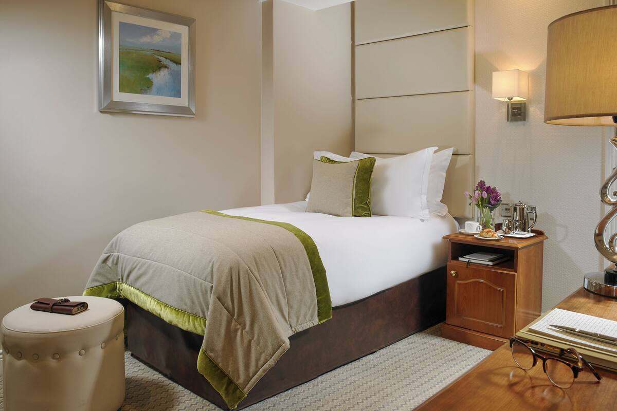 Una habitación de hotel ordenada con cama, mesita de noche, lámpara y un taburete pequeño.