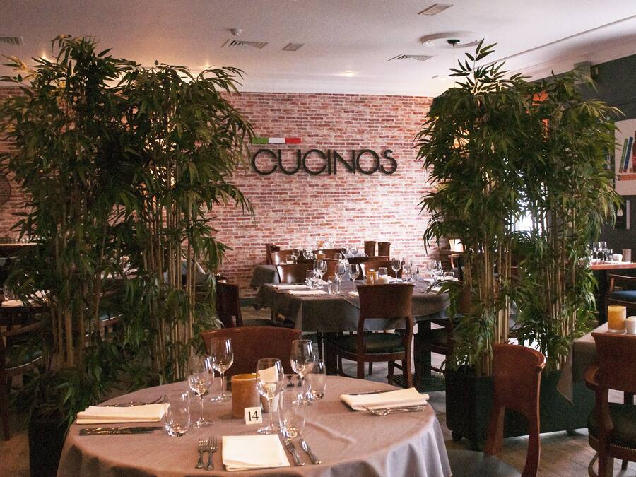 A restaurant interior with brick walls, tables set with wine glasses and cutlery, and potted plants.