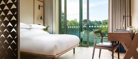 Habitación de hotel con una cama grande, un escritorio con silla, una planta en maceta y vistas a zonas verdes y edificios a través de la ventana.