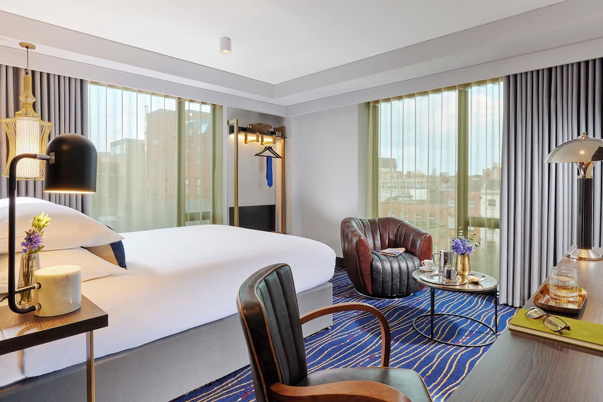 A modern hotel room with a large bed, a leather armchair, a small table with a tray, and a desk with a lamp and vase.